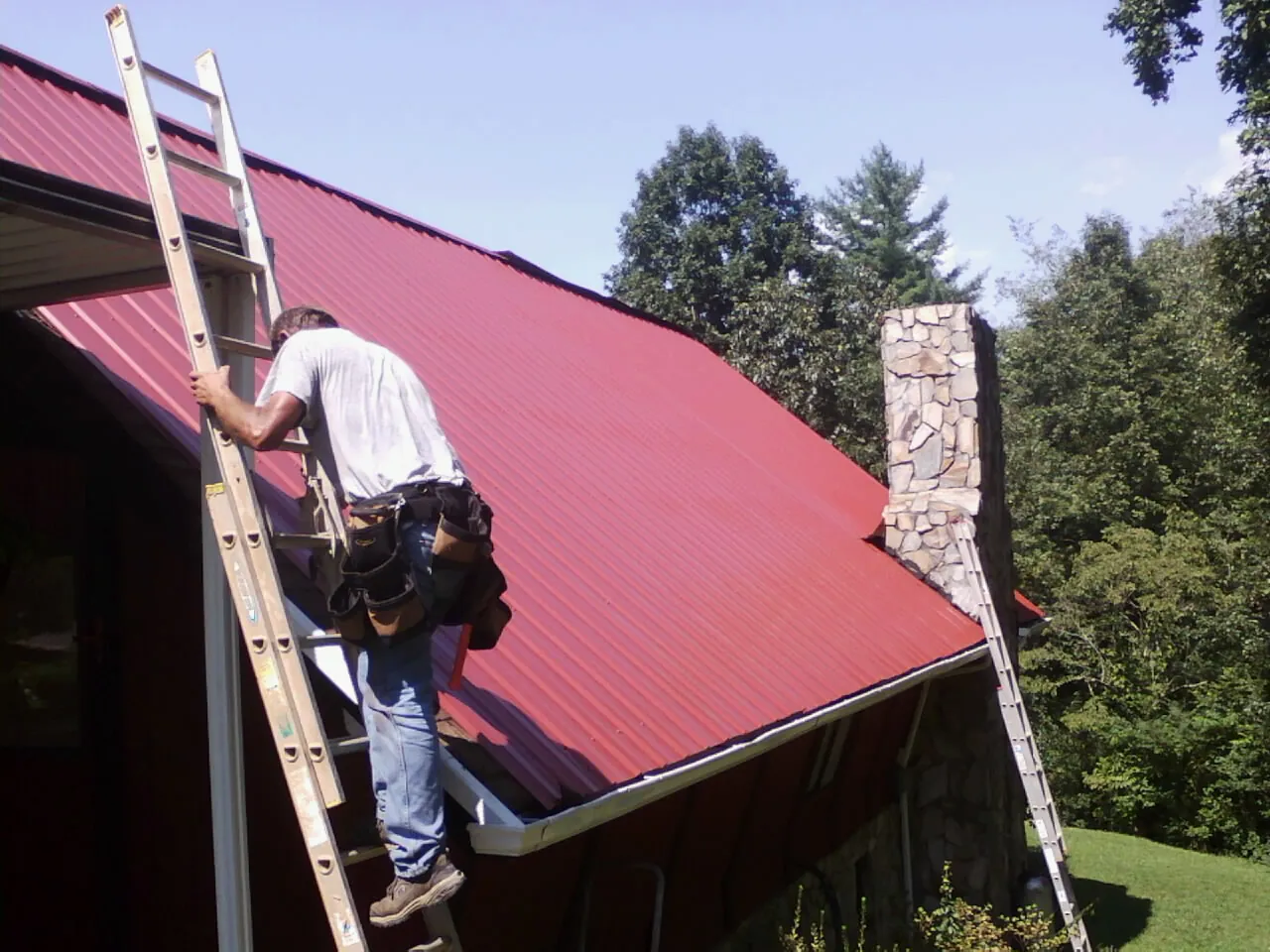 Roofer on a ladder beside a red metal roof and stone chimney — Soffit & Fascia Repair by Roofing Brothers in Trail